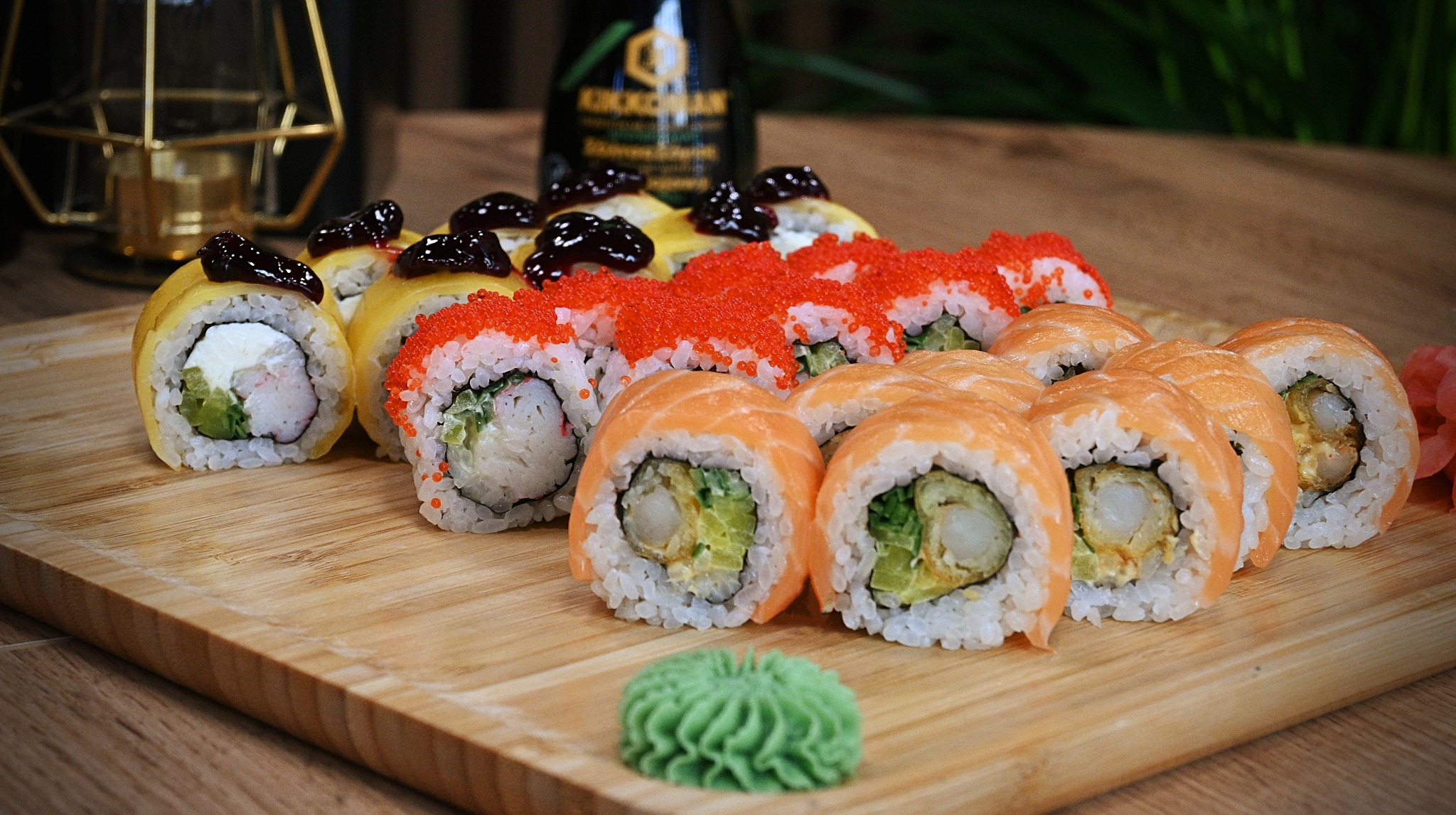 Różnorodne sushi na drewnianej desce: rolki z łososiem, kawiorem i omletem japońskim, w tle butelka sosu sojowego i dekoracyjny świecznik geometryczny.
