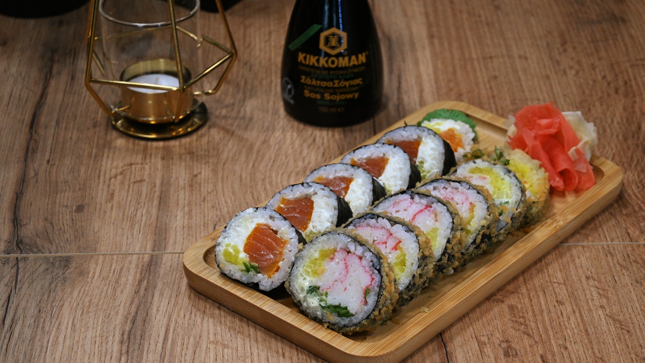 Różnorodny zestaw sushi na bambusowej tacy, z imbirem i butelką sosu sojowego Kikkoman w tle, obok dekoracyjnej świeczki w geometrycznym świeczniku.