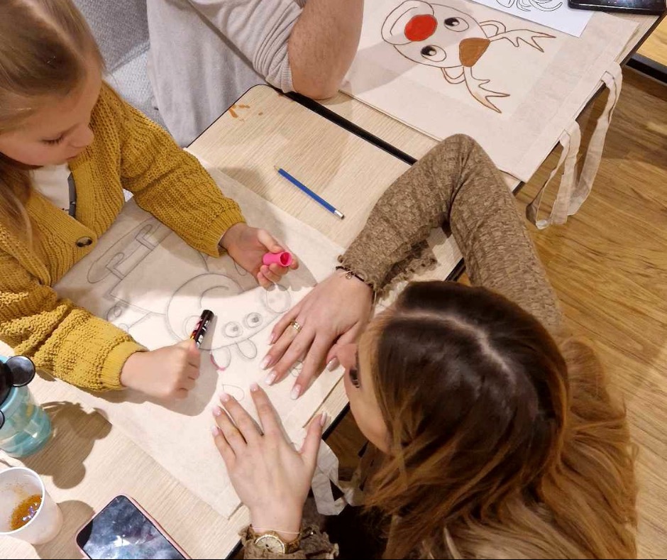 Dziecko i dorosły dekorują bawełniane torby markerami na stole. Na torbach widoczne szkice reniferów. Kreatywne warsztaty dla dzieci.