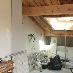 Blackwings - Poddasze w trakcie remontu: drewniana boazeria na suficie, nieotynkowana ściana z widocznymi przewodami, sterta desek i wiórów na podłodze, drabina i narzędzia w tle.