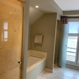 Blackwings - Wykończona łazienka z beżowymi płytkami imitującymi marmur na podłodze i ścianach, przeszklona kabina prysznicowa z beżowego kamienia, wanna obudowana płytkami, okno z żaluzjami.