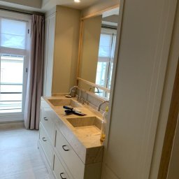 Blackwings - Wykończona łazienka z umywalką z beżowego marmuru, białe szafki z czarnymi uchwytami, fragment okna z zasłoną, na umywalce leżą narzędzia.