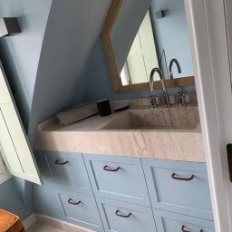 Blackwings - Łazienka na poddaszu ze skośnym sufitem, jasnoniebieskie szafki z beżowym blatem i umywalką z kamienia, lustro w drewnianej ramie, nowoczesna armatura i otwarte okienko.