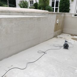 Blackwings - Szlifowanie ścian basenu zewnętrznego przed wykończeniem, widoczna szlifierka z podłączonym odkurzaczem przemysłowym, surowe ściany, schody do basenu w tle.