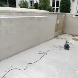 Szlifowanie ścian basenu zewnętrznego przed wykończeniem, widoczna szlifierka z podłączonym odkurzaczem przemysłowym, surowe ściany, schody do basenu w tle.