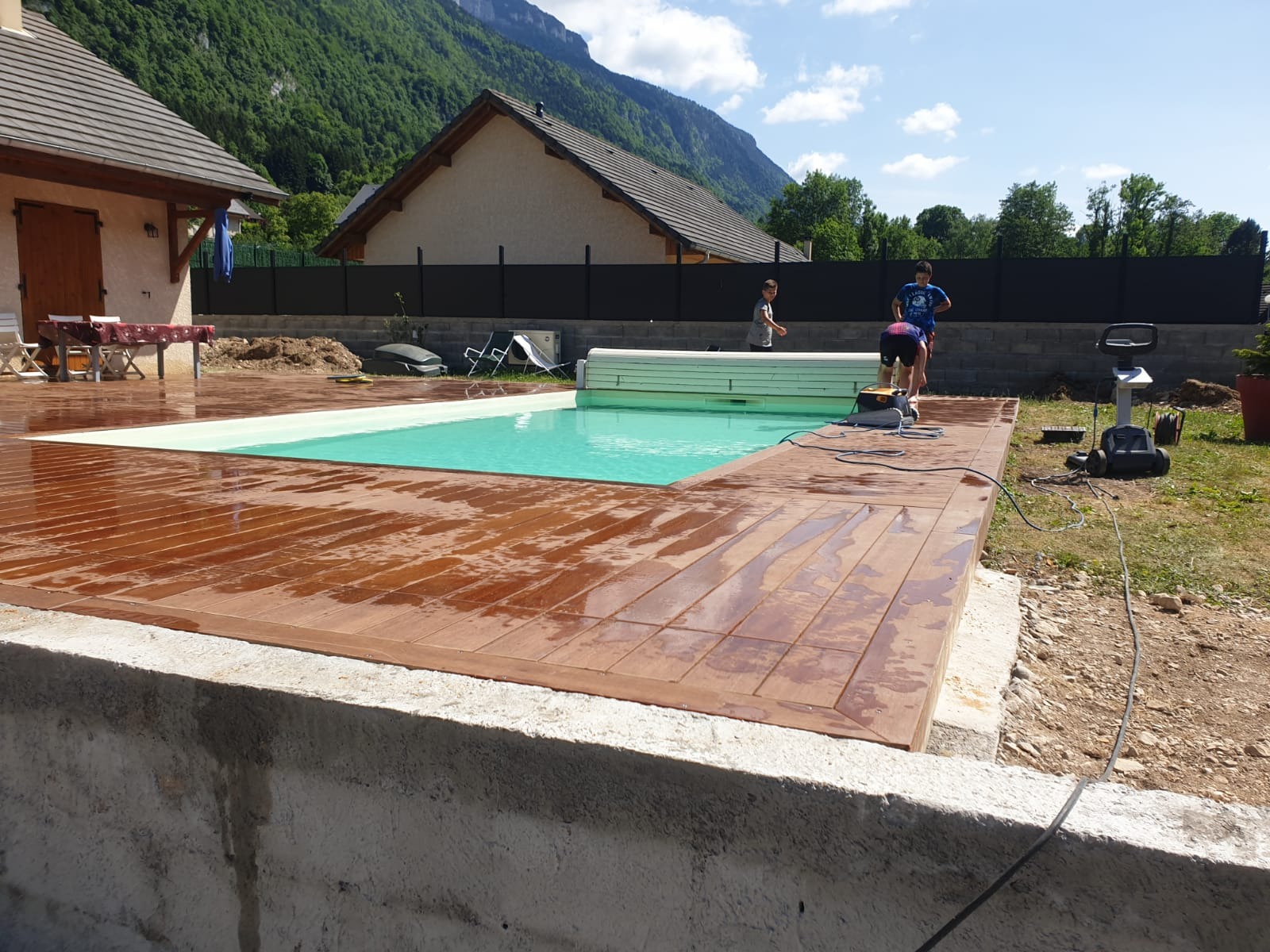 Nowy, drewniany taras wokół basenu w ogrodzie, z widokiem na góry w tle, w trakcie czyszczenia przez dwóch chłopców, obok stoi urządzenie czyszczące.
