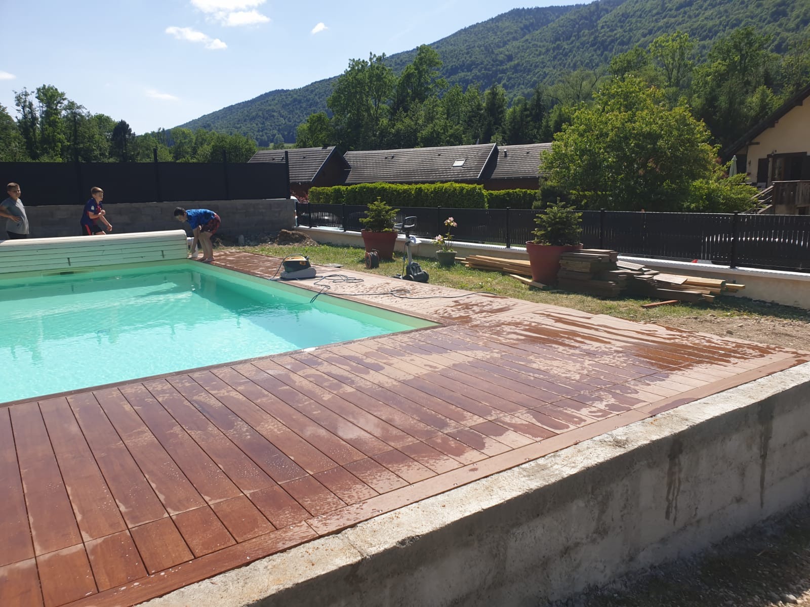 Montaż drewnianego tarasu wokół basenu zewnętrznego, widoczne narzędzia, deski i osoby w tle, zieleń i góry w oddali.