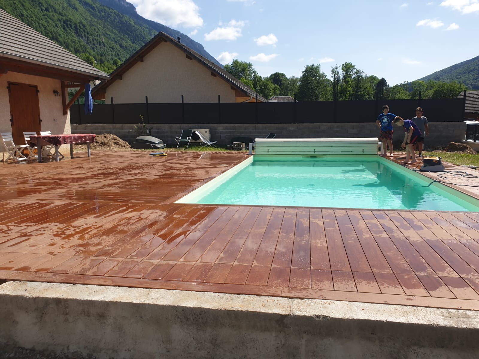 Nowo wybudowany basen z drewnianym tarasem wokół, w tle góry i domy, kilku chłopców stoi przy basenie, prawdopodobnie sprawdzając jakość wody.