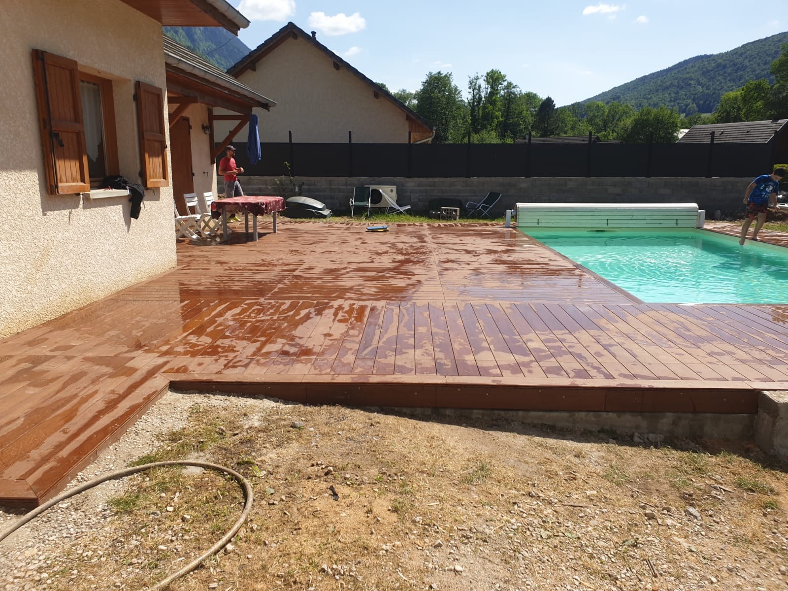 Świeżo wyczyszczony, brązowy taras z drewna kompozytowego przylegający do jasnego domu, obok basenu z turkusową wodą, w tle góry i domy.