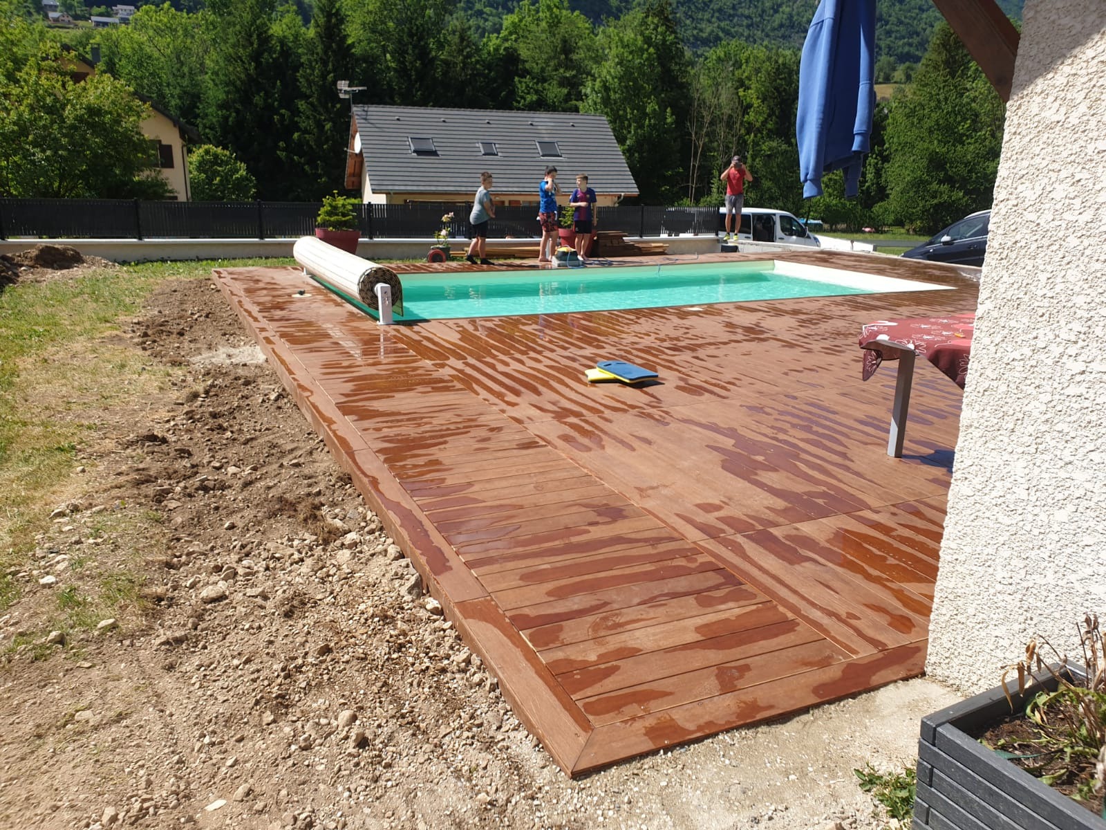 Nowa drewniana podłoga wokół basenu ogrodowego, częściowo mokra po czyszczeniu, z widocznym zwiniętym zabezpieczeniem basenu i grupą osób w tle.