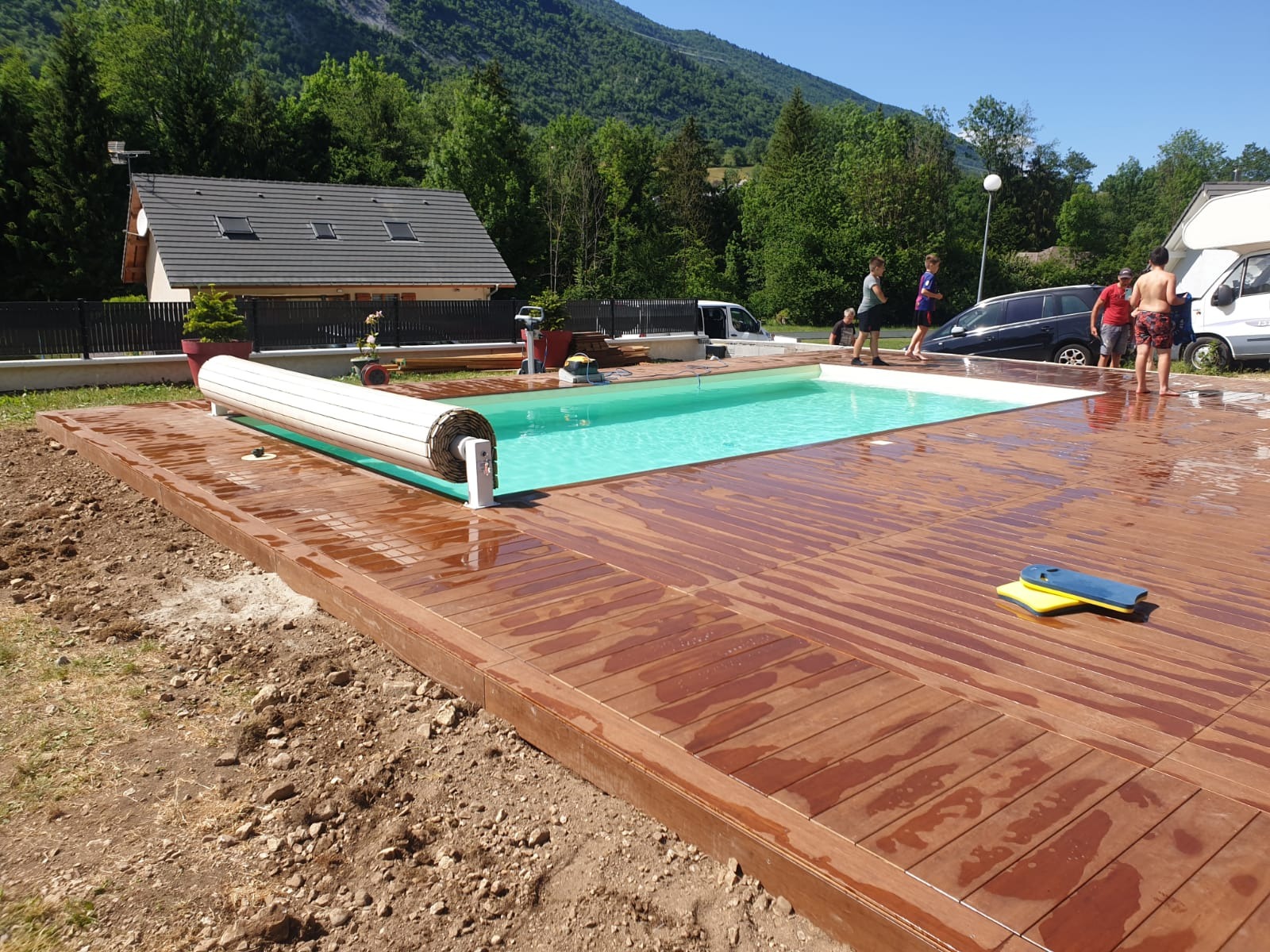 Nowoczesny basen z drewnianym tarasem w trakcie budowy, widoczny zwinięty roleta basenowa, w tle dom i góry.