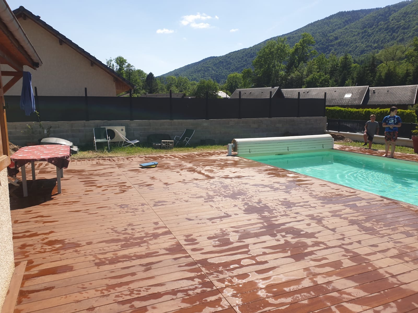 Świeżo wyczyszczony drewniany taras przy basenie w ogrodzie z widokiem na góry w słoneczny dzień. Widoczne ślady wody na deskach i częściowo zwinięta roleta basenowa.