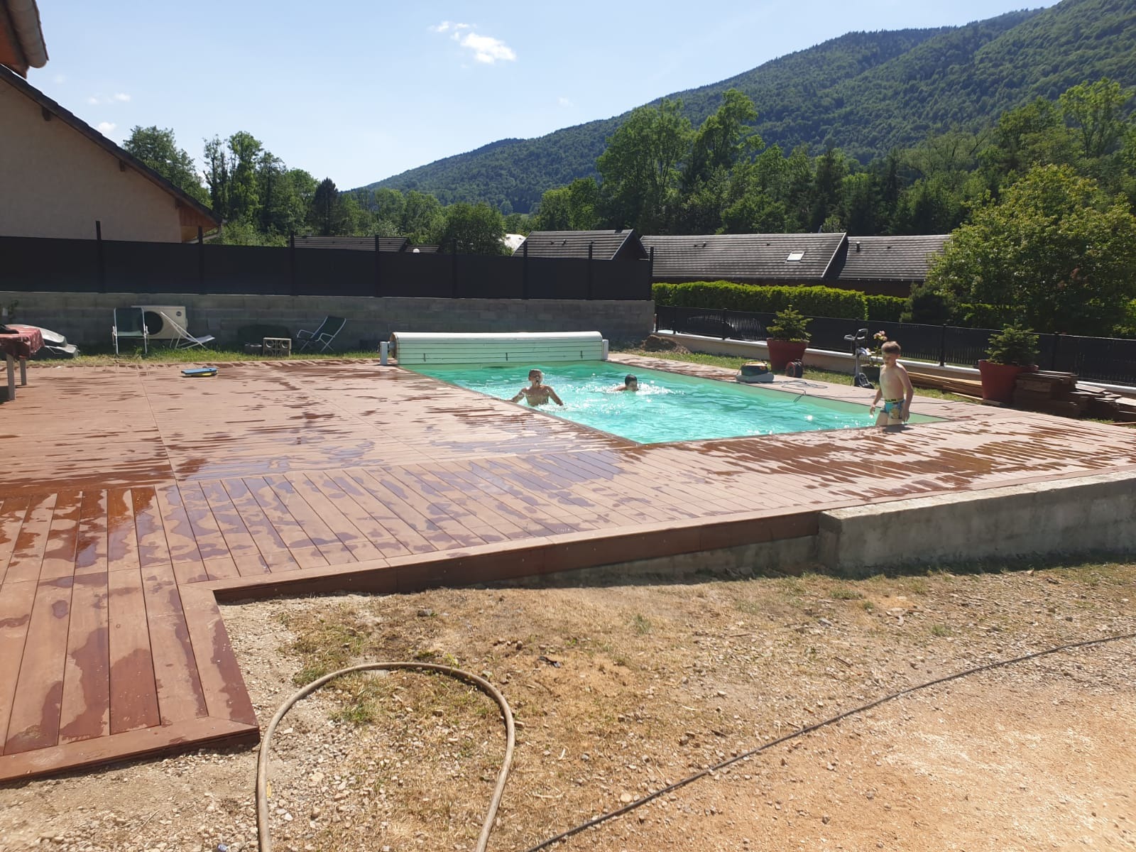 Drewniany taras okalający basen z dwójką dzieci pływających i jednym wychodzącym z wody, na tle zieleni i gór.