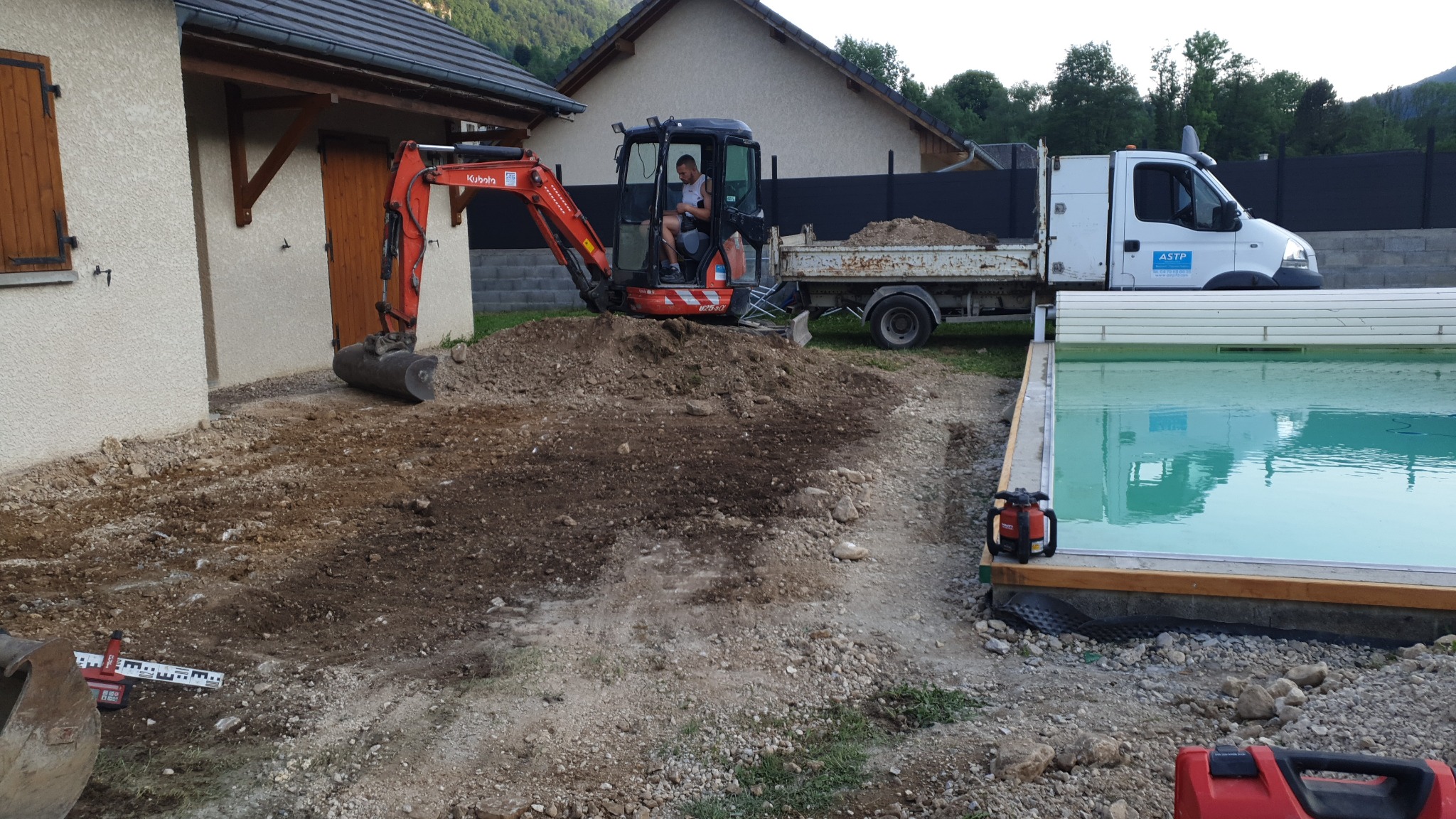 Przygotowanie terenu pod budowę basenu: minikoparka Kubota i wywrotka na działce z domem w tle.