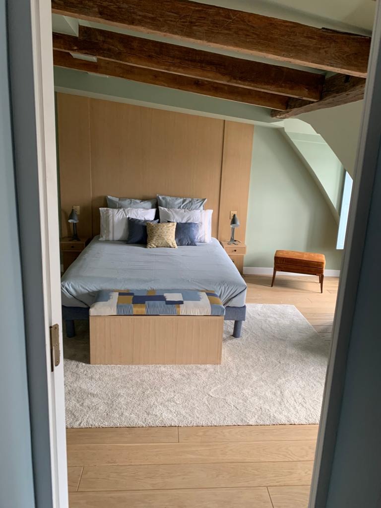 Sypialnia w odcieniach beżu i błękitu z drewnianym stropem, widoczna przez otwarte drzwi, z łóżkiem z wezgłowiem z paneli, pufą w patchworkowy wzór i dywanem z długim włosiem.