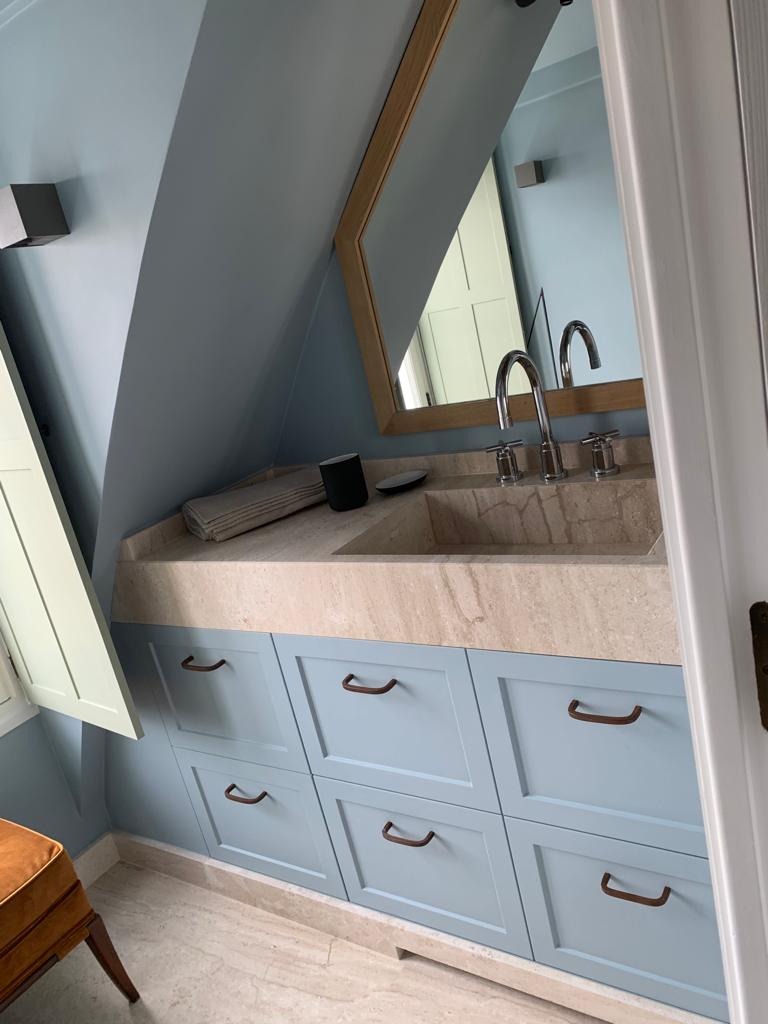 Łazienka na poddaszu ze skośnym sufitem, jasnoniebieskie szafki z beżowym blatem i umywalką z kamienia, lustro w drewnianej ramie, nowoczesna armatura i otwarte okienko.