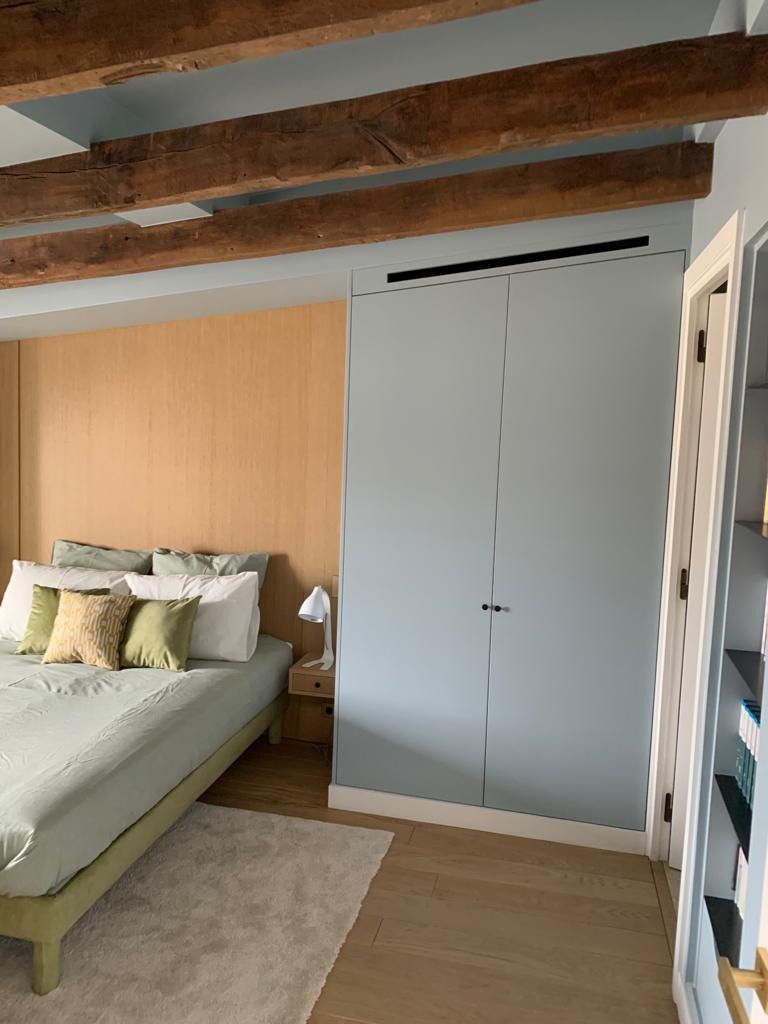 Jasna sypialnia z drewnianymi belkami stropowymi, zabudowaną szafą w kolorze pastelowego błękitu, łóżkiem z zieloną pościelą i drewnianą ścianą za wezgłowiem.