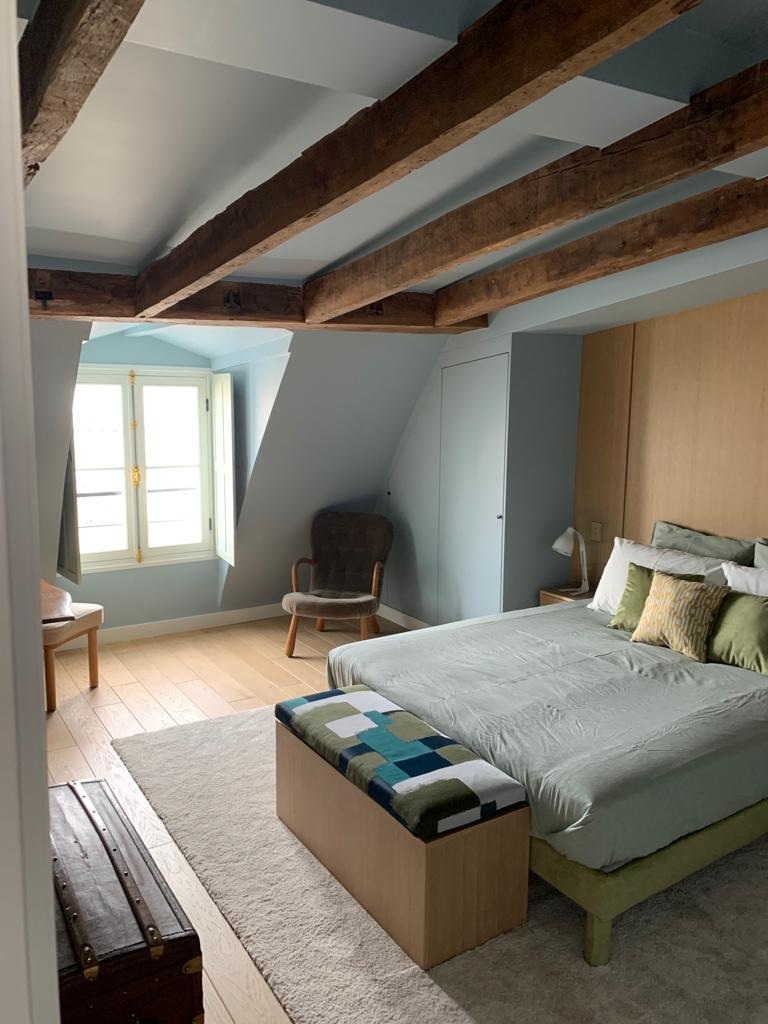Jasna sypialnia na poddaszu z drewnianymi belkami stropowymi, beżowym dywanem, łóżkiem z zieloną narzutą i skrzynią obitą tkaniną w geometryczne wzory.