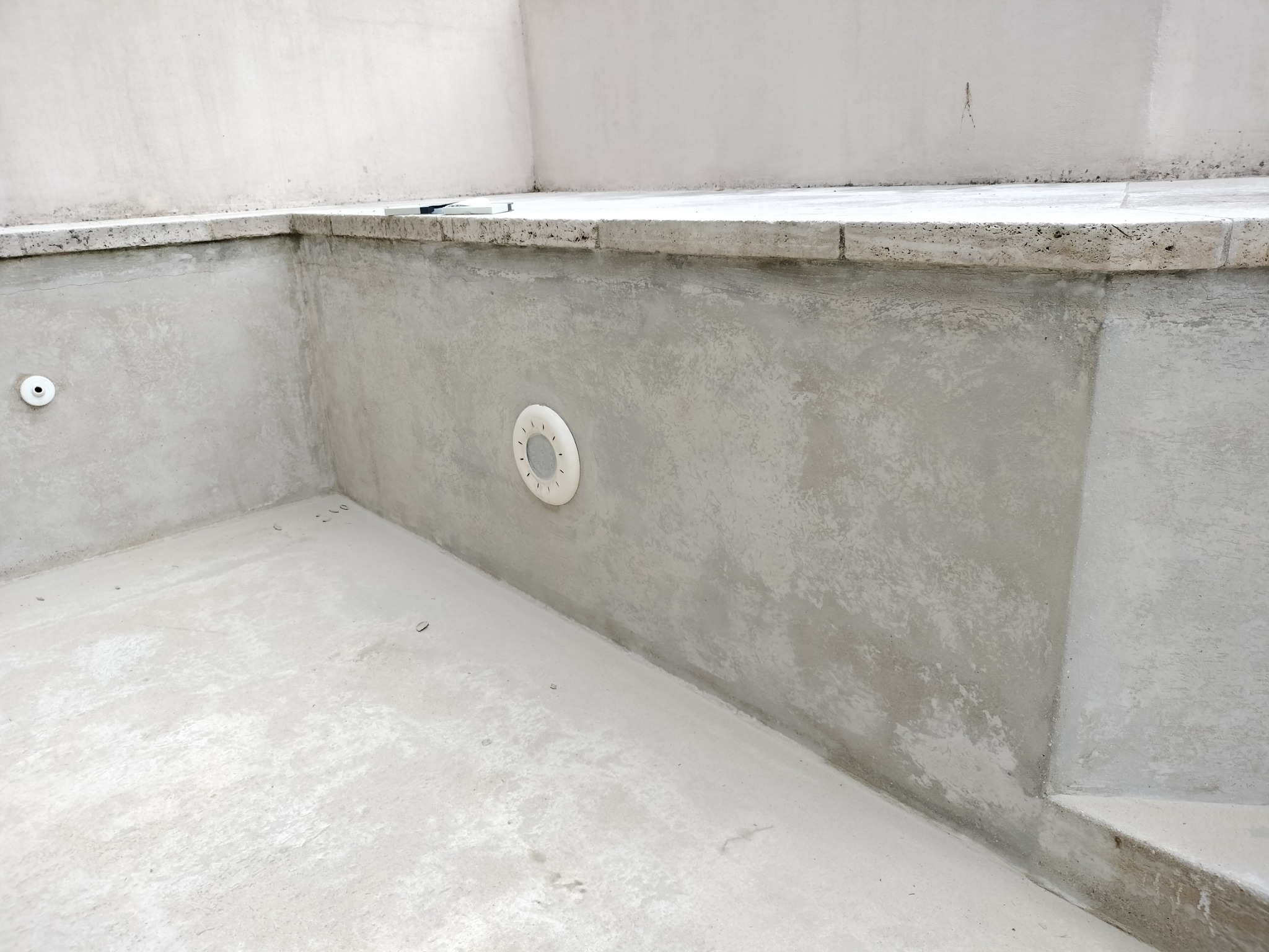 Wnętrze pustego basenu w trakcie budowy lub remontu, szare ściany i dno, widoczny odpływ w ścianie i stopnie.