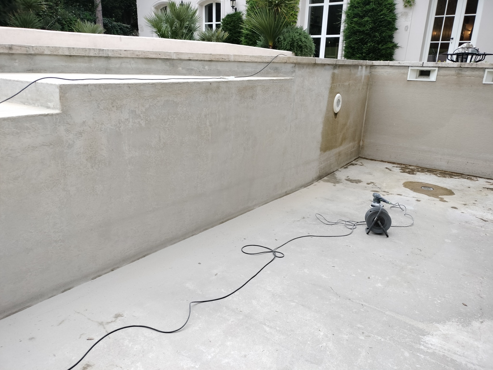 Szlifowanie ścian basenu zewnętrznego przed wykończeniem, widoczna szlifierka z podłączonym odkurzaczem przemysłowym, surowe ściany, schody do basenu w tle.