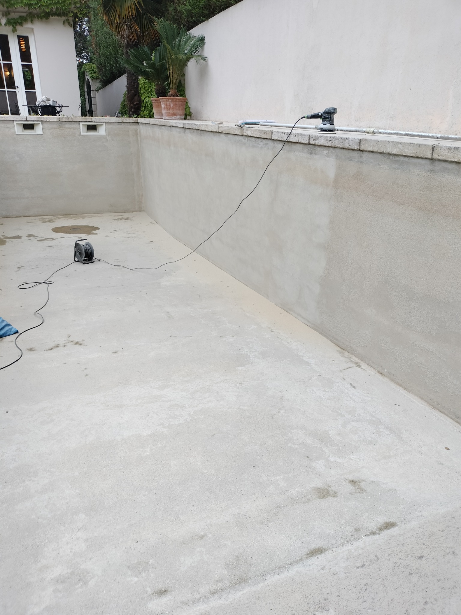 Wnętrze pustego basenu w trakcie renowacji, widoczna szlifierka na krawędzi i zwinięty przedłużacz na dnie, ściany basenu w surowym betonie.