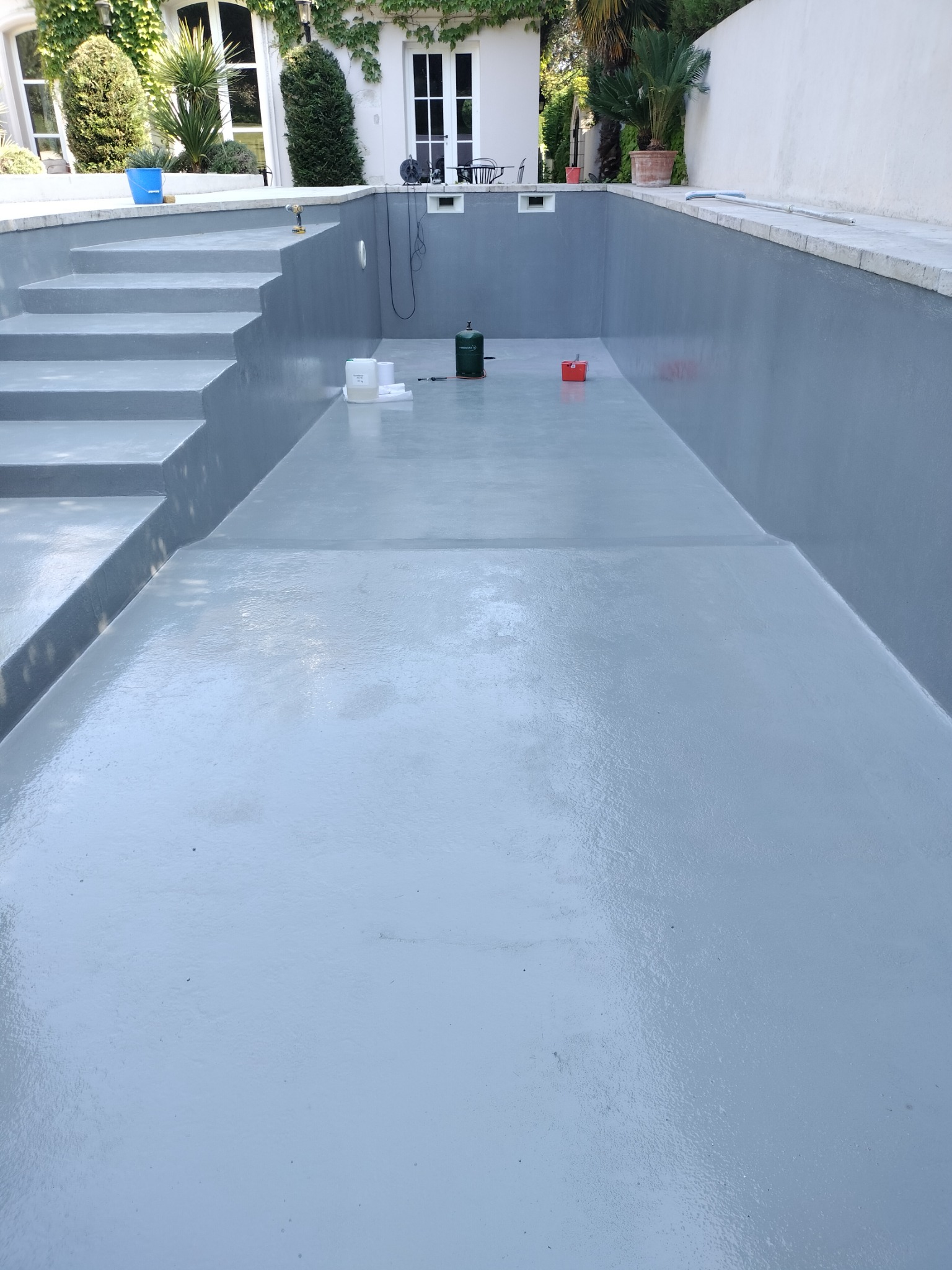 Świeżo pomalowany, pusty basen ogrodowy w odcieniach szarości, z widocznymi schodami wejściowymi i akcesoriami malarskimi na dnie.