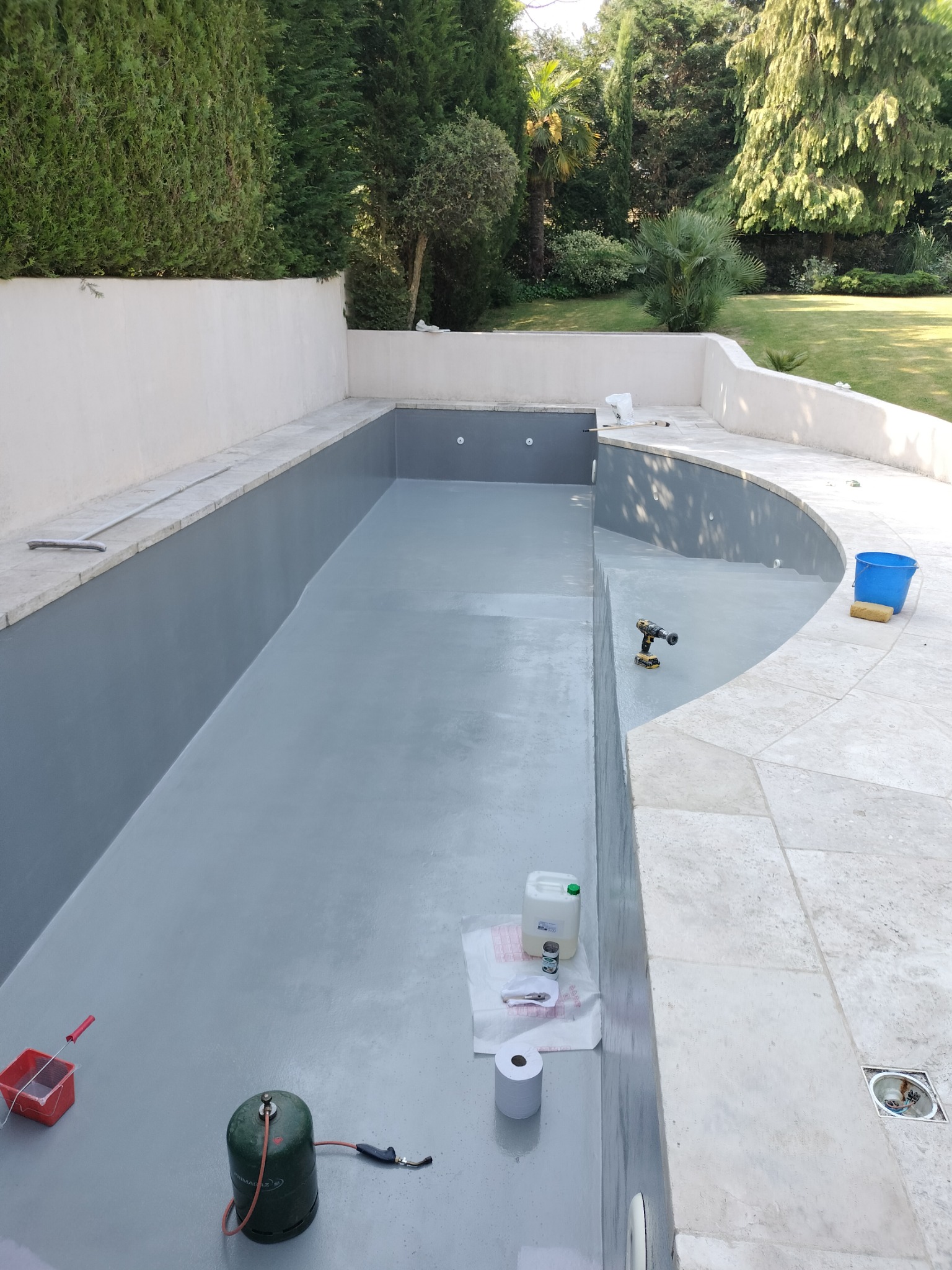 Szary basen w trakcie malowania, widoczne narzędzia: butla gazowa, pędzel, wiadro z farbą, wiertarka, pojemnik z płynem, rolka papieru. W tle zadbany ogród z drzewami.