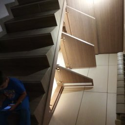 Meble Agart Artur Cichoń - Schody z drewnianymi stopniami i wbudowanymi szafkami z jasnego drewna, z widocznymi zawiasami i szufladami, dziecko siedzi na schodach i używa telefonu.