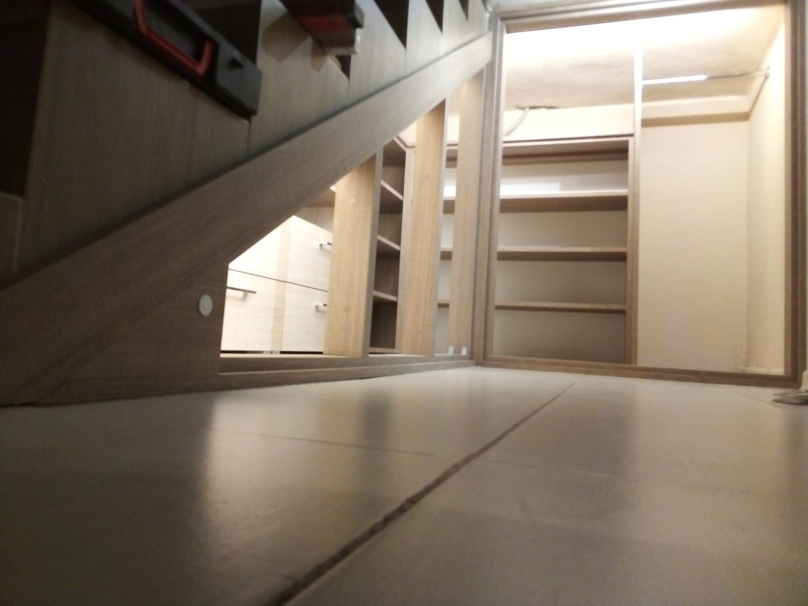 Niski kąt ujęcia prezentuje przestrzeń pod schodami z zabudową meblową: szuflady i regał z półkami oraz szafa wnękowa z oświetleniem wewnętrznym.