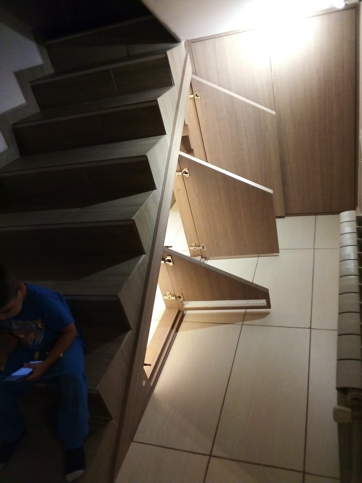 Schody z drewnianymi stopniami i wbudowanymi szafkami z jasnego drewna, z widocznymi zawiasami i szufladami, dziecko siedzi na schodach i używa telefonu.