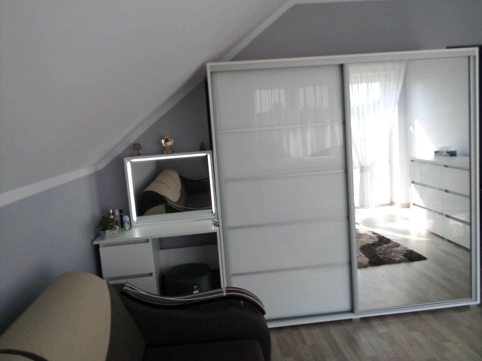 Biała szafa przesuwna z lustrem w pokoju na poddaszu, obok toaletka z podświetlanym lustrem i pamiątki na blacie, widoczny fragment sofy.
