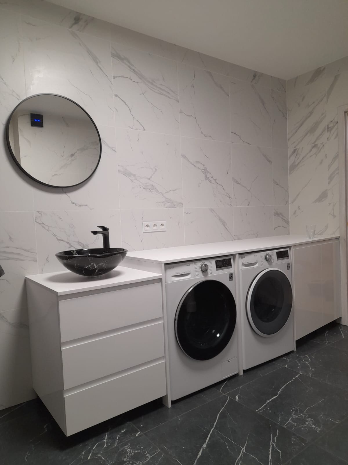 Nowoczesna łazienka z marmurowymi płytkami, czarną umywalką w kształcie misy na białej szafce z szufladami oraz dwiema pralkami pod blatem.