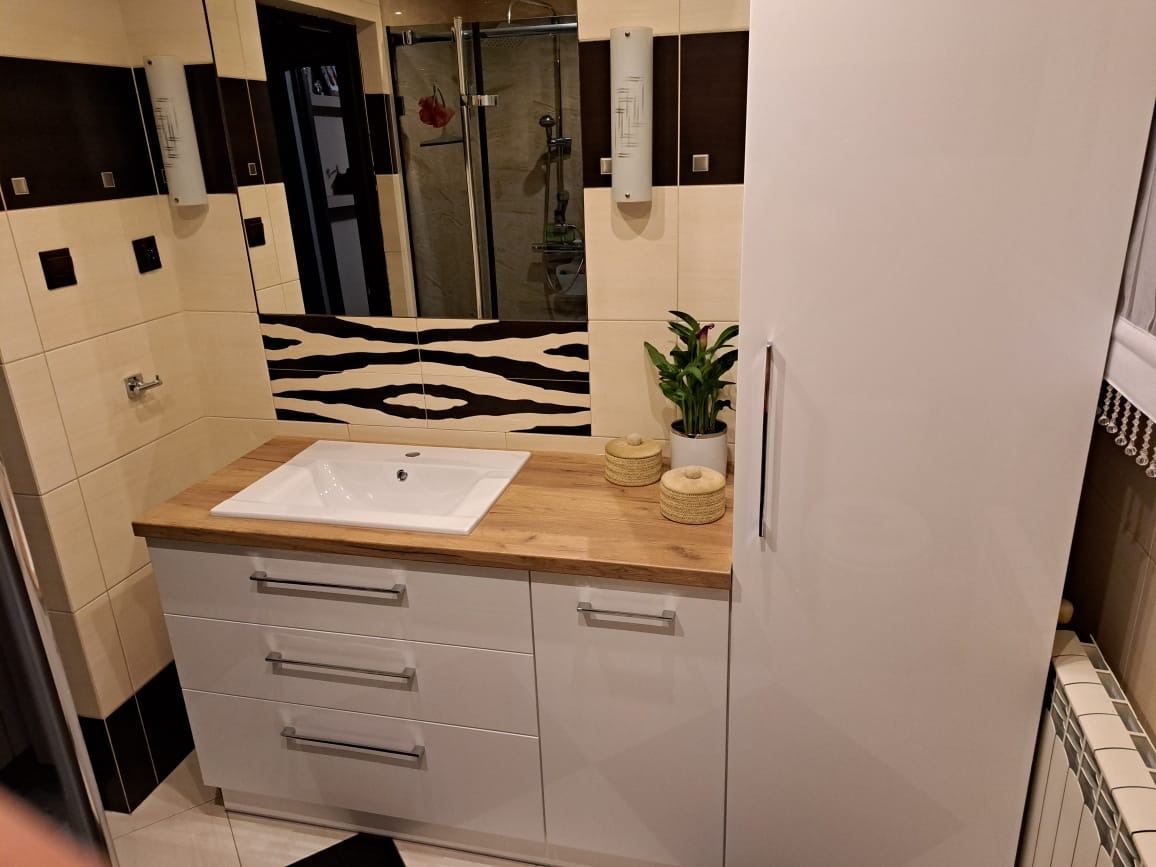 Łazienka z białą szafką pod umywalką z drewnianym blatem, nad nią lustro z czarno-białymi wzorami zebry na ścianie, obok szafka z rośliną doniczkową i ozdobnymi pojemnikami.