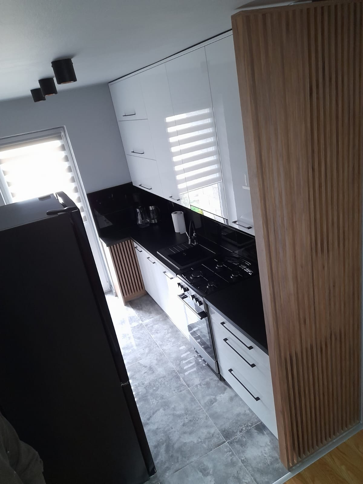 Nowoczesna kuchnia z białymi szafkami, czarnym blatem i drewnianą ścianką działową, widok z góry na płytki podłogowe.