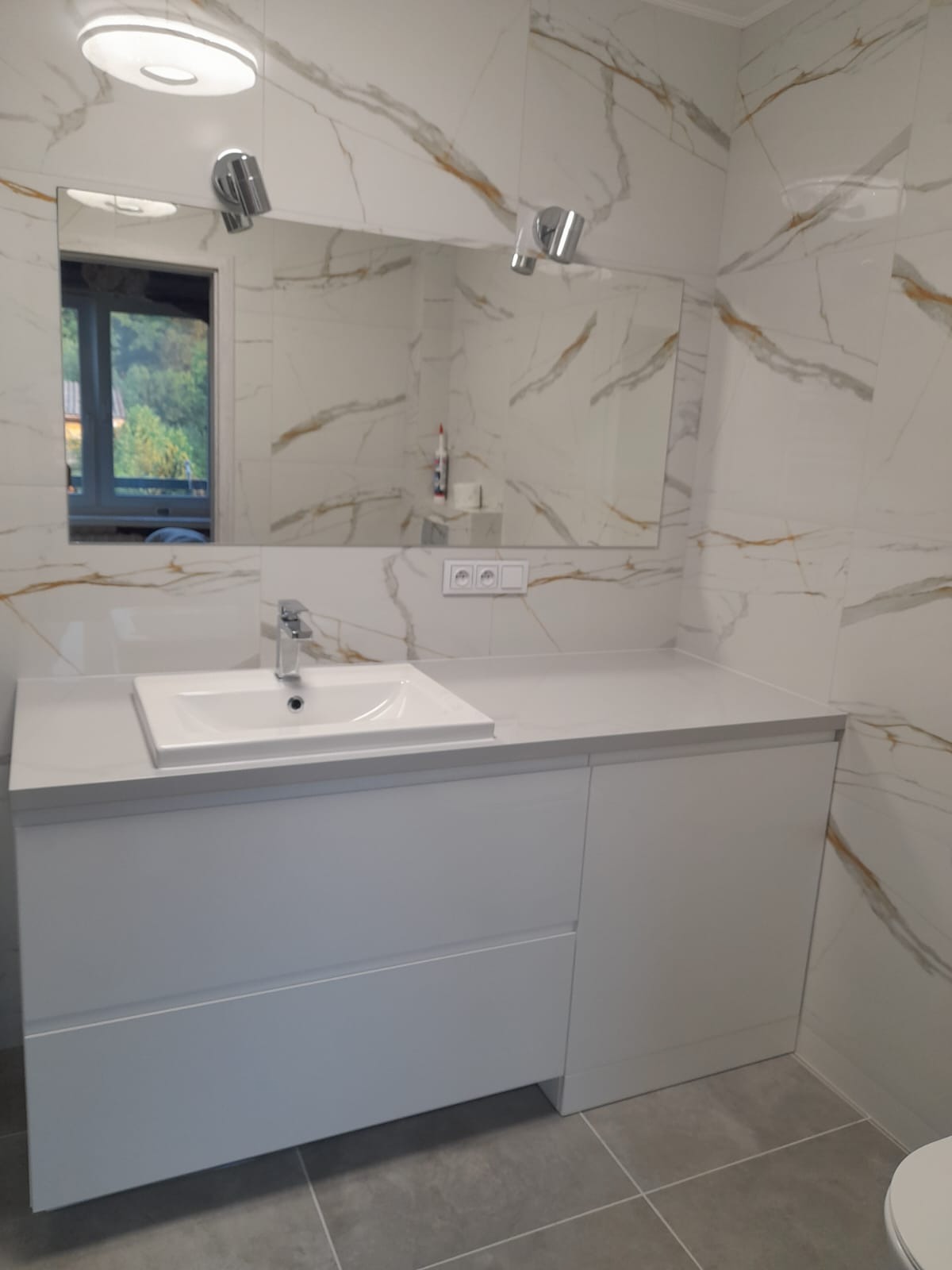 Biała umywalka nablatowa z szafką pod umywalkową i baterią, zamontowana na tle ściany wyłożonej płytkami imitującymi marmur z złotymi żyłami, oświetlona lampkami punktowymi.