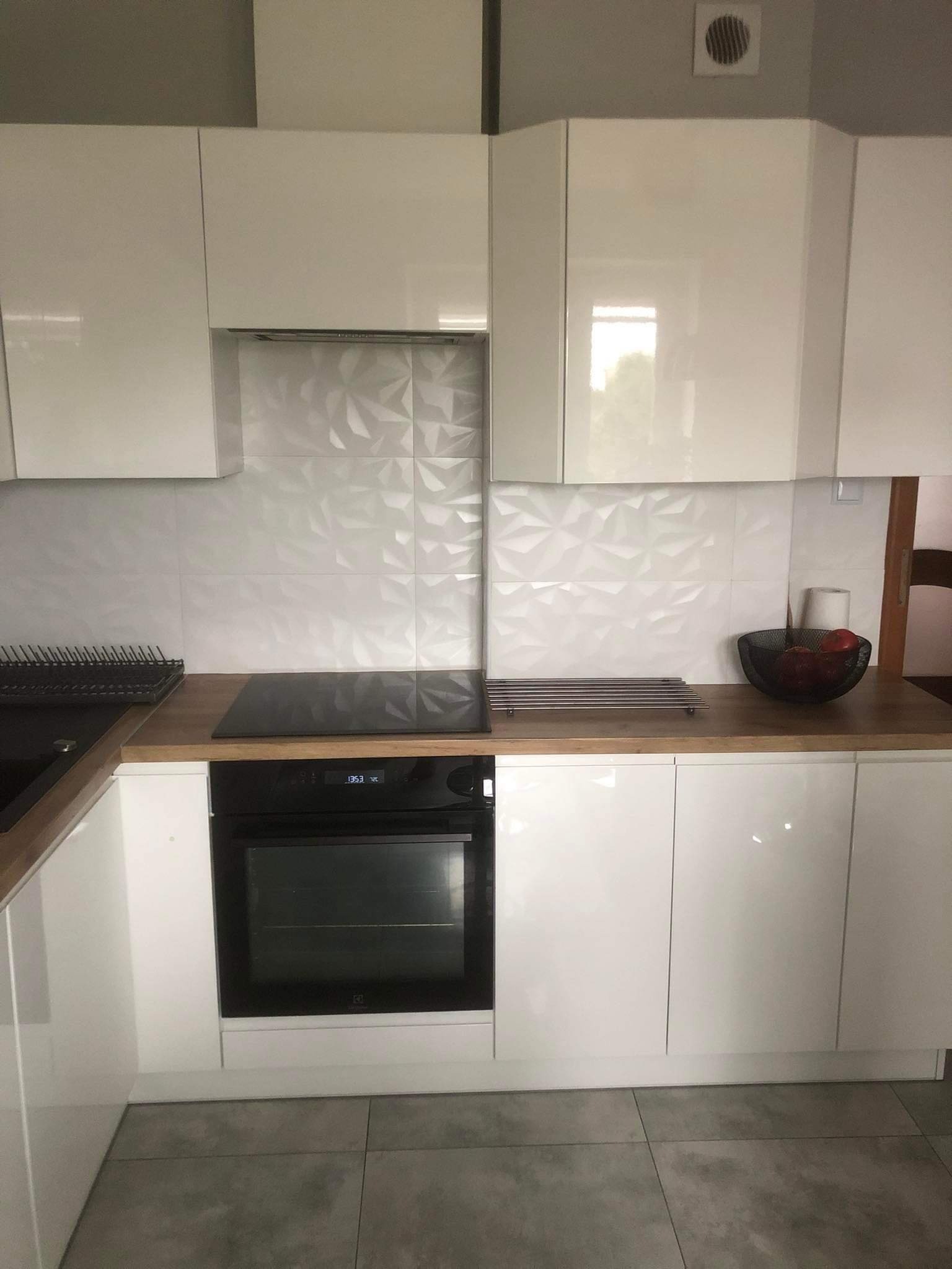 Biała kuchnia z lakierowanymi frontami, drewnianym blatem, płytą indukcyjną i piekarnikiem, ozdobne białe płytki na ścianie.