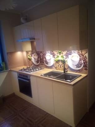 Jasna zabudowa kuchenna z blatem drewnopodobnym i panelem dekoracyjnym z motywem kawy, widoczny zlew dwukomorowy i płyta gazowa.