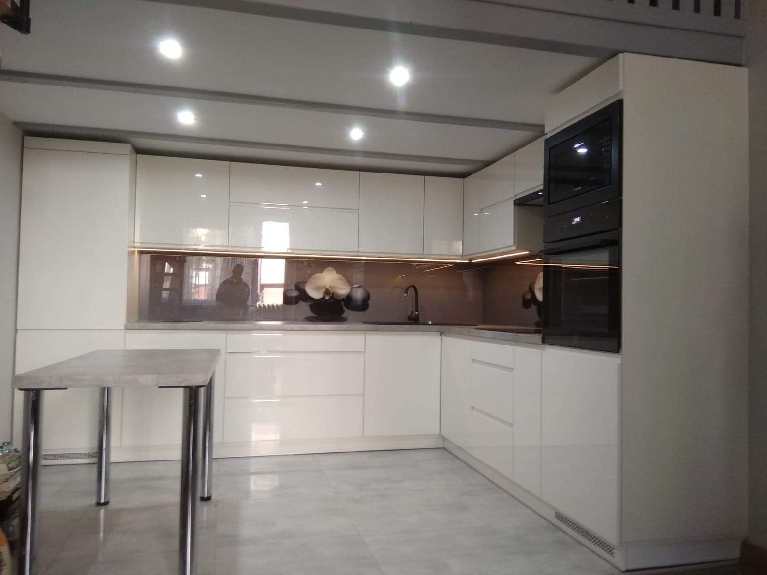 Nowoczesna kuchnia z białymi, lakierowanymi frontami, podświetleniem LED pod szafkami wiszącymi i ciemnym panelem szklanym nad blatem, na którym stoi wazon z orchideą. Na blacie widoczne odbicie...