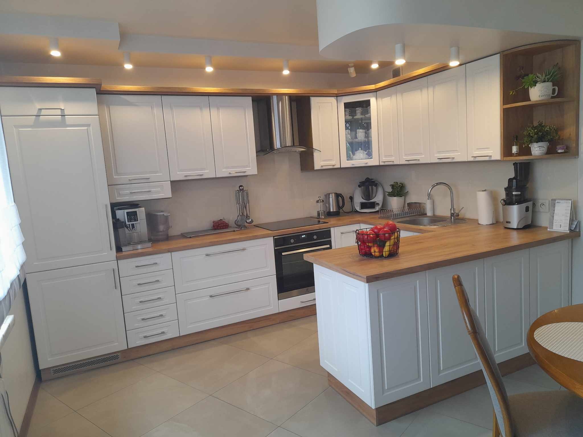 Jasna, przestronna kuchnia z białymi szafkami, drewnianymi blatami i oświetleniem punktowym, widoczne owoce w koszyku i elementy dekoracyjne.