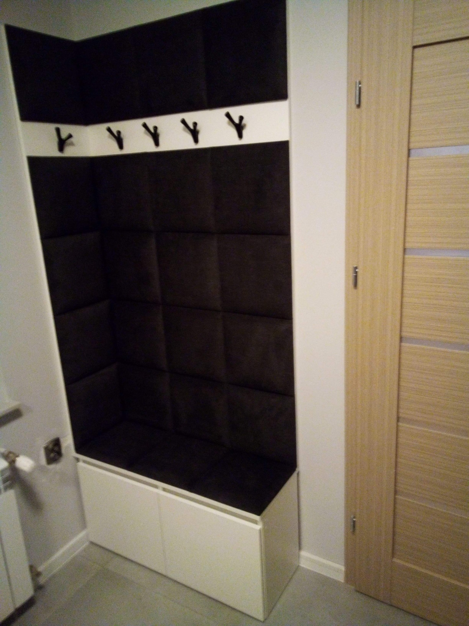 Wnęka w przedpokoju z miękkim, pikowanym obiciem w kolorze ciemnego brązu, biała listwa z haczykami na ubrania i szafka z siedziskiem w kolorze białym, obok drewniane drzwi.