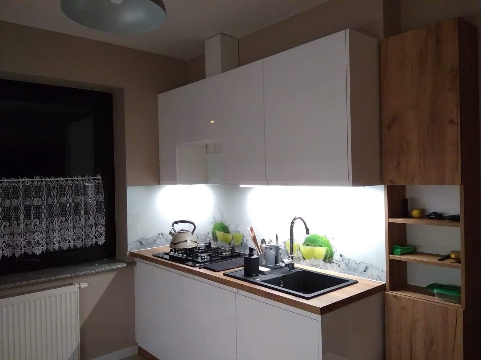 Nowoczesna kuchnia z białymi szafkami, drewnianym blatem i szklanym panelem nad blatem z motywem cytryn, widok na czarny zlew i płytę grzewczą.