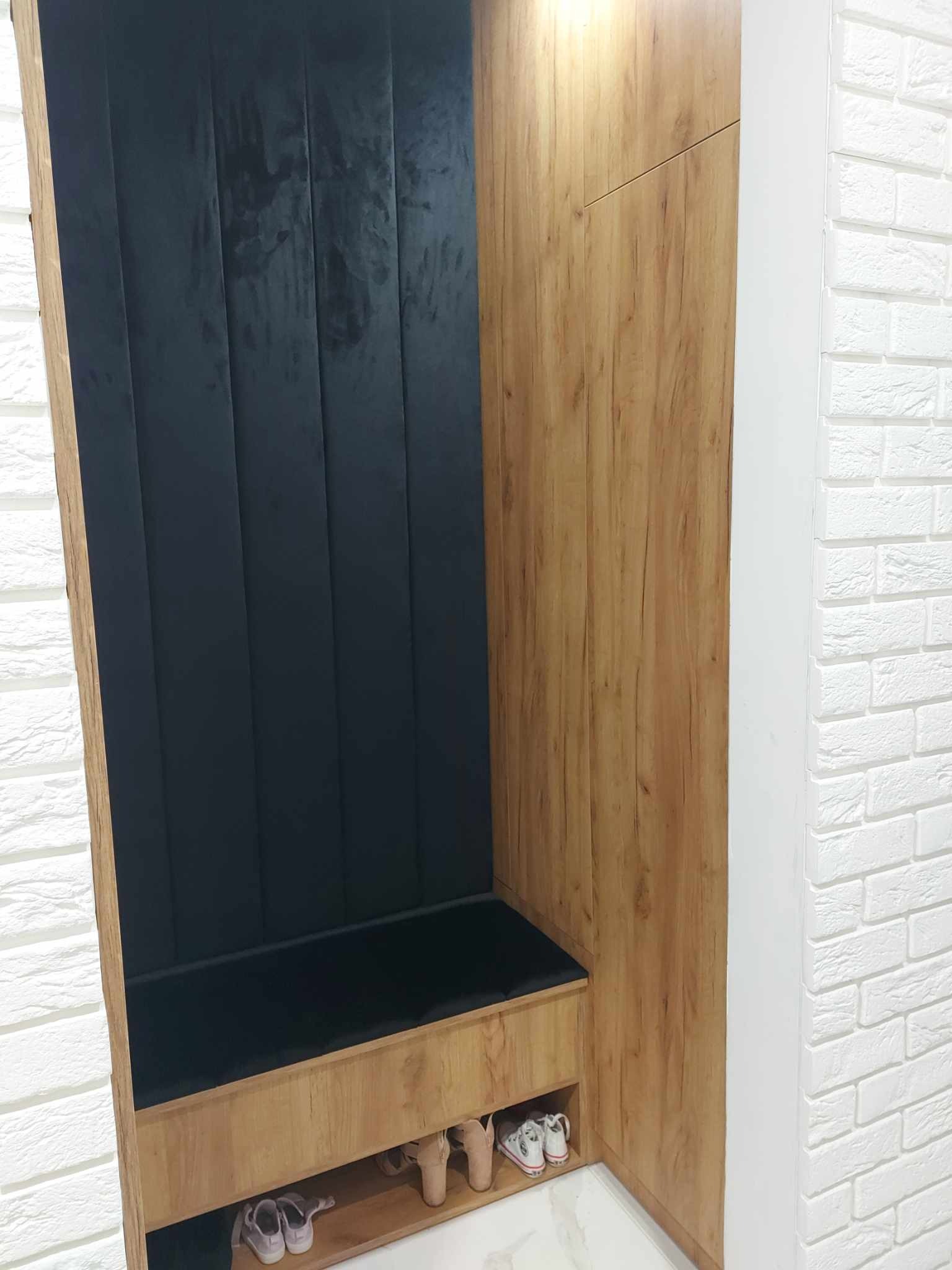 Wnęka z siedziskiem i panelem tapicerowanym w kolorze ciemnego granatu, szafa z okleiną drewnopodobną, biała ściana z dekoracyjnej cegły, buty w dolnej półce.