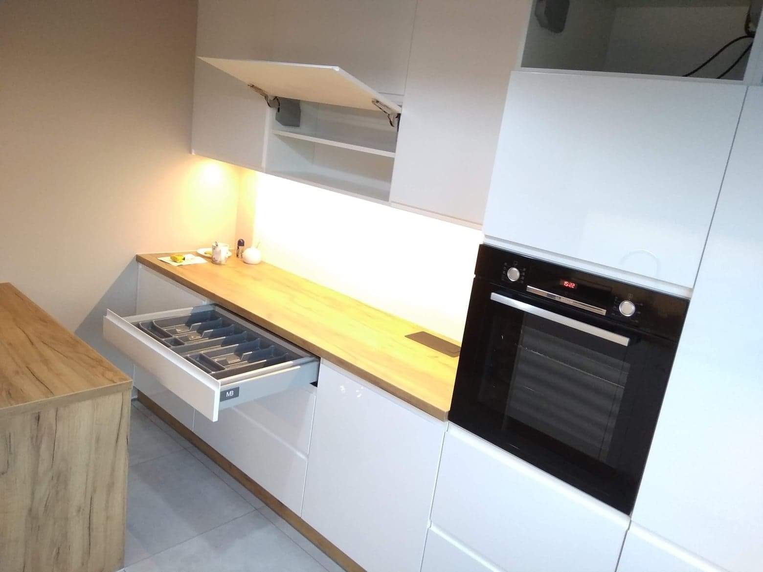 Nowoczesna biała zabudowa kuchenna z drewnianym blatem, wysunięta szuflada z organizerem na sztućce, piekarnik w zabudowie i szafka otwierana do góry.