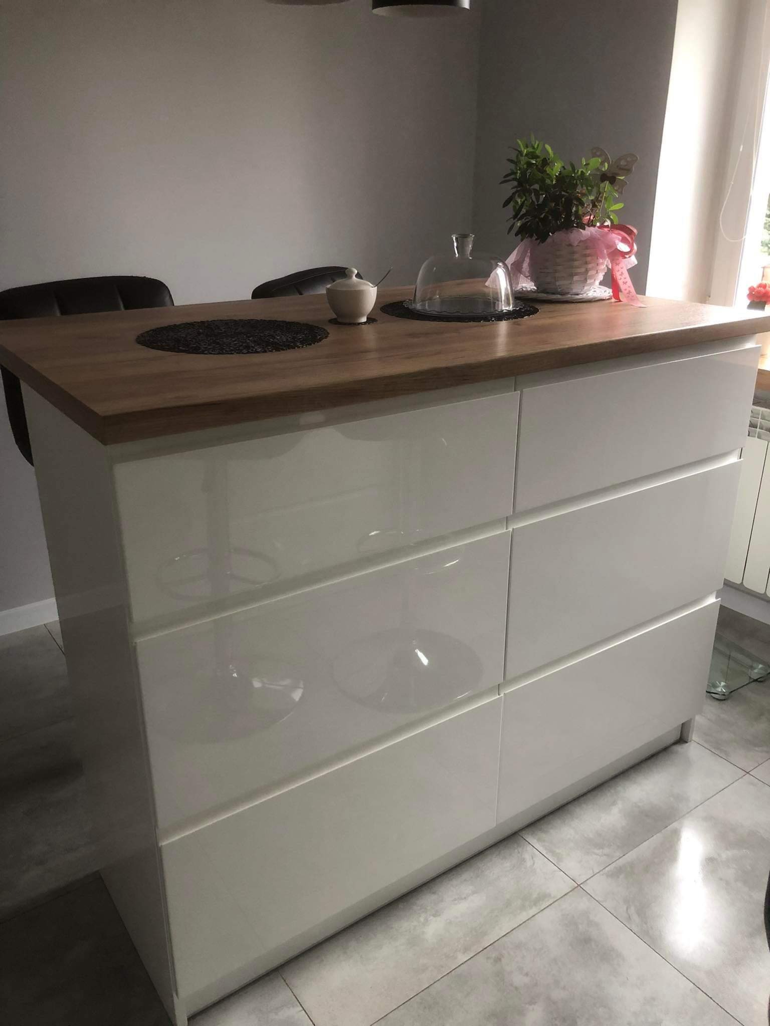 Wyspa kuchenna z białymi, lakierowanymi frontami szuflad i drewnianym blatem, na którym stoi roślina w ozdobnym koszyku i szklany klosz. W tle widoczne krzesła barowe i okno.