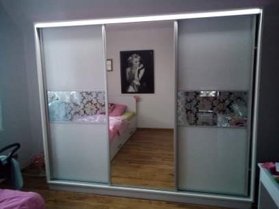 Biała szafa przesuwna z lustrzanymi drzwiami i dekoracyjnym pasem z motywem kwiatowym, odbijająca wnętrze pokoju z łóżkiem i obrazem Marilyn Monroe na ścianie.