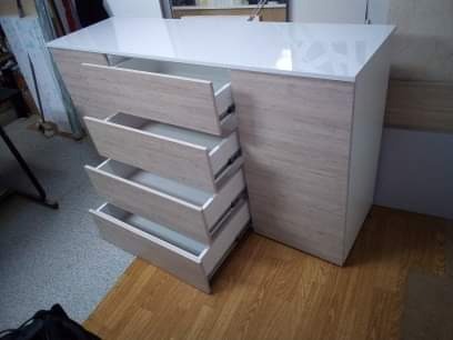 Komoda z czterema wysuniętymi szufladami o jasnym, drewnopodobnym wykończeniu, z białym, błyszczącym blatem, ustawiona na drewnianej podłodze, widoczna z perspektywy lekko z góry.