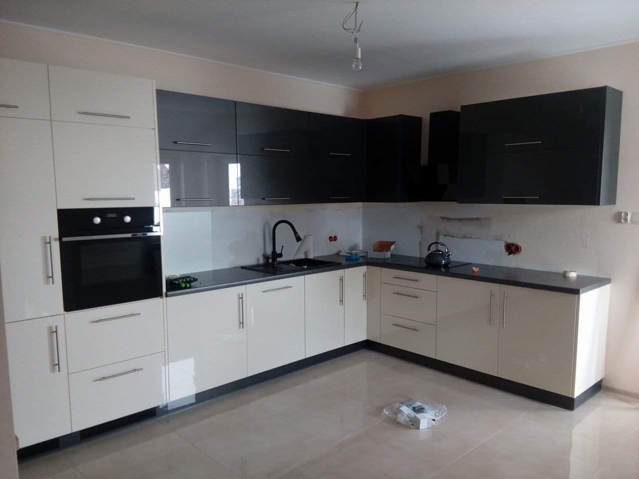 Nowoczesna kuchnia z białymi i grafitowymi szafkami z połyskiem, czarnym blatem i zlewozmywakiem, widok na fragment ściany w trakcie wykończenia w Nowym Broniszewie.