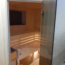 Jasne drewno i szklane drzwi w saunie z ławkami, widok z zewnątrz. Minimalistyczny design, ciepłe oświetlenie, widoczny piec i kratka wentylacyjna.