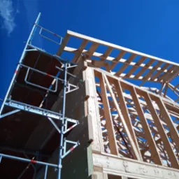 Wznoszenie drewnianej konstrukcji szkieletowej budynku w Zubrzycy Dolnej, widok od dołu na rusztowanie i elementy konstrukcyjne na tle błękitnego nieba.