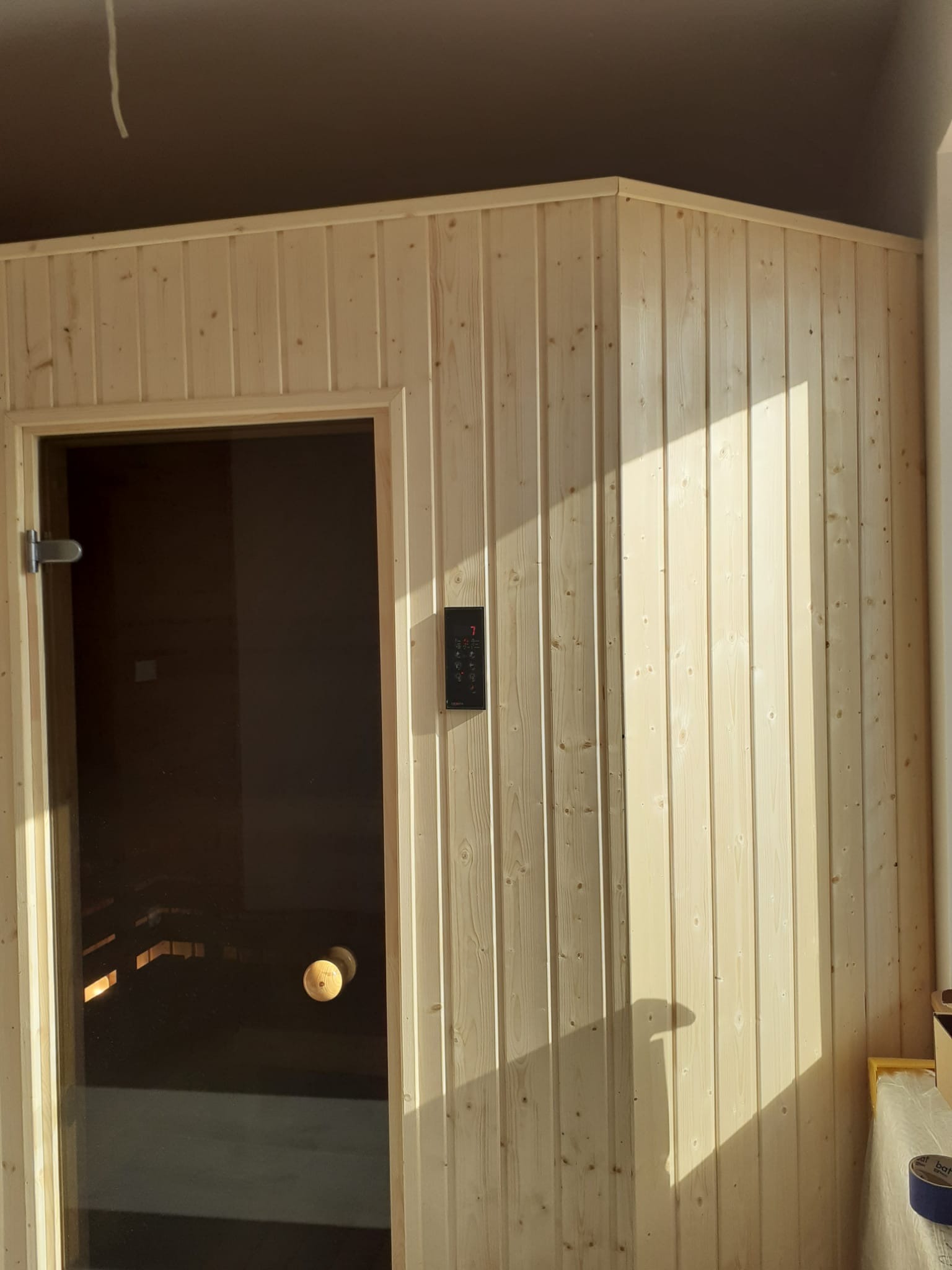 Jasna, drewniana sauna z przeszklonymi drzwiami i panelem sterowania. Widoczne pionowe deski i naturalne oświetlenie.